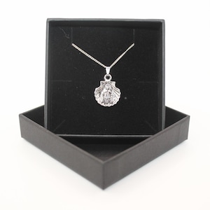 May include: A silver pendant necklace with a religious figure in a shell design, presented in an open black gift box. The necklace has a delicate silver chain. The box has a black velvet interior.