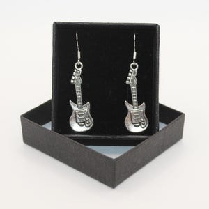 Puede incluir: Un par de pendientes de guitarra plateados en una caja de joyería de terciopelo negro.