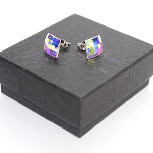 May include: A pair of square stud earrings featuring a multi-faceted, iridescent surface that reflects shades of blue, purple, and yellow. The earrings have a metallic post backing and are displayed on a textured black square jewellery box.