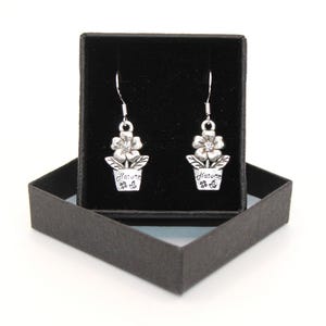 May include: A pair of silver-toned flower earrings with a small clear gem in the center of each flower. The earrings are in a black gift box. Each earring has the word "Nature" and a small flower design on the pot.