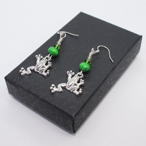 May include: A pair of silver frog earrings with green glass beads. The earrings are displayed on a black box.