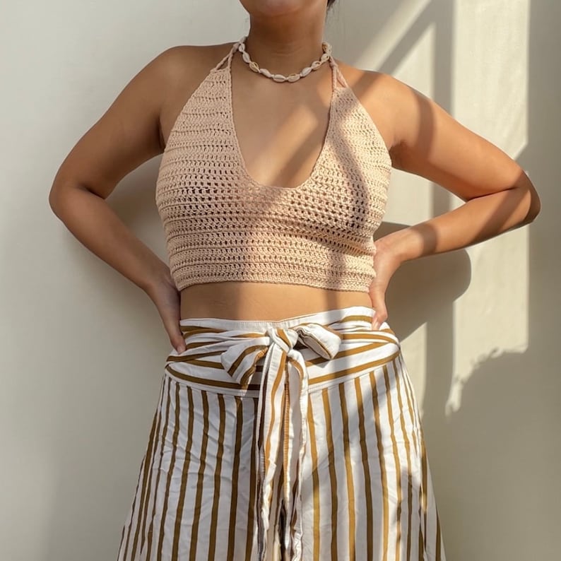 Puede incluir: Un top halter de crochet rosa claro con un collar de conchas. El top se combina con pantalones de rayas blancas y marrones.