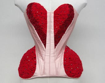 May include: A pale pink corset with red heart-shaped accents, embellished with small, sparkling details. The corset is displayed on a white mannequin. The text "ARMANDO GABRIEL COUTURE CORSETRY" is visible at the bottom.