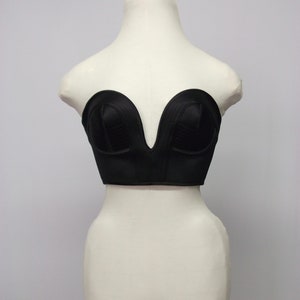 Pode incluir: Um top de corset preto com decote em V profundo, exibido em um manequim branco. O corset apresenta painéis estruturados e um acabamento liso, semelhante ao cetim. O texto "ARMANDO GABRIEL COUTURE CORSETRY" está impresso no topo.