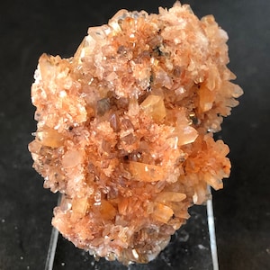 May include: A cluster of orange and white crystals on a clear display stand. The crystals are arranged in a random pattern and have a rough, textured surface.