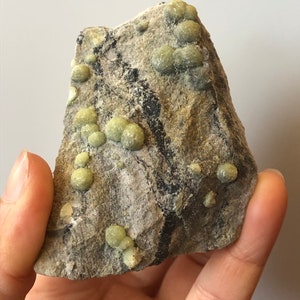 Puede incluir: Una roca gris rugosa con grupos de pequeños cristales redondos verdes. La roca tiene un aspecto natural y terroso.
