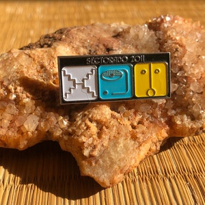 Puede incluir: Un pin de metal plateado con tres cuadrados. El primer cuadrado es blanco con un patrón blanco y negro. El segundo cuadrado es azul con un círculo blanco y líneas negras. El tercer cuadrado es amarillo con un círculo negro y dos líneas negras. El pin dice "Sectorado 2011".