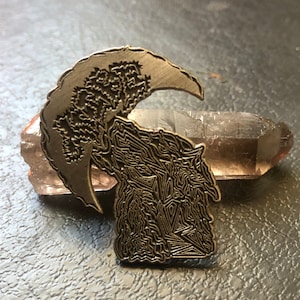 Puede incluir: Un pin de metal plateado con un diseño detallado de una luna creciente y un árbol. El pin está sobre una superficie marrón.