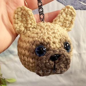 Puede incluir: Un llavero de ganchillo hecho a mano con la cabeza de un bulldog francés de color canela con ojos azules y una nariz negra. El llavero tiene un cierre y una cadena de metal oscuro. El texto "woodlandsandwhimsy" es visible en la parte inferior.