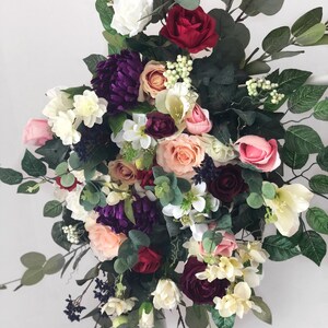 May include: A lush floral arrangement featuring a variety of flowers in shades of pink, red, white, and purple. The arrangement includes roses, hydrangeas, and other blooms, as well as greenery.