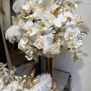 K&ouml;nnte beinhalten: Ein wei&szlig;-goldener Blumenstrau&szlig; mit wei&szlig;en Orchideen, Rosen und Federn in einer goldenen Vase.
