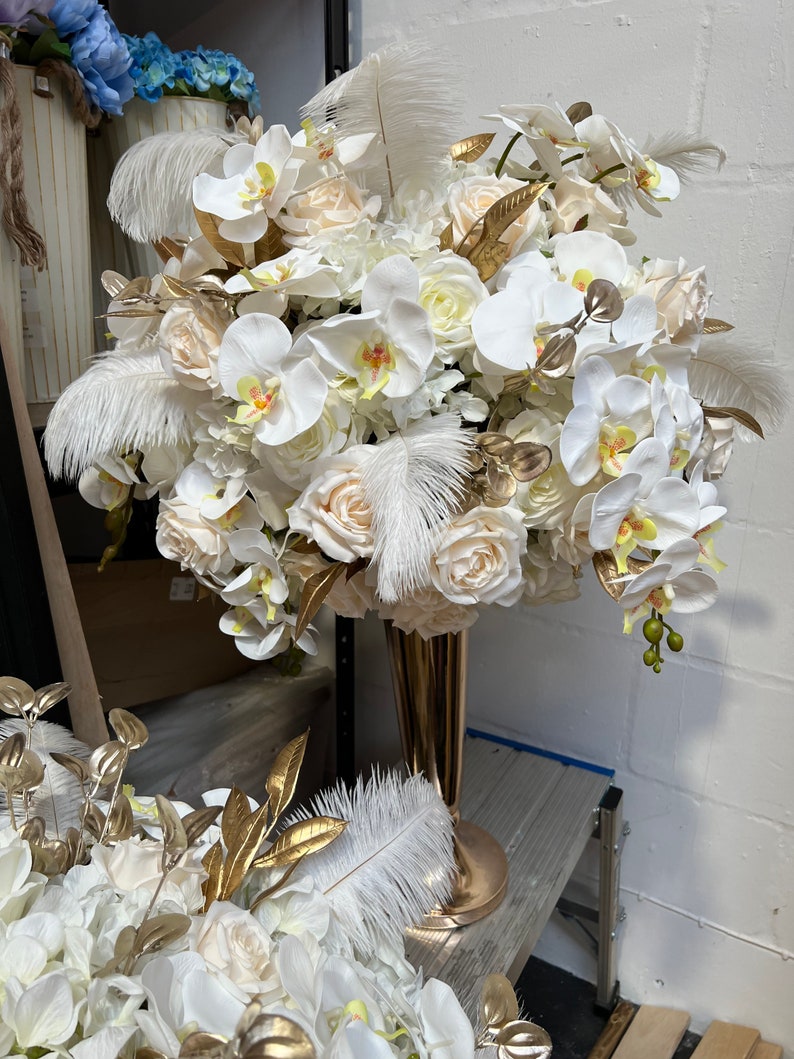 K&ouml;nnte beinhalten: Ein wei&szlig;-goldener Blumenstrau&szlig; mit wei&szlig;en Orchideen, Rosen und Federn in einer goldenen Vase.