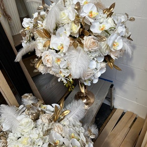 K&ouml;nnte beinhalten: Ein wei&szlig;-goldener Blumenstrau&szlig; mit wei&szlig;en Orchideen, Rosen und Hortensien. Die Anordnung befindet sich in einer goldenen Vase und ist mit wei&szlig;en Federn verziert.