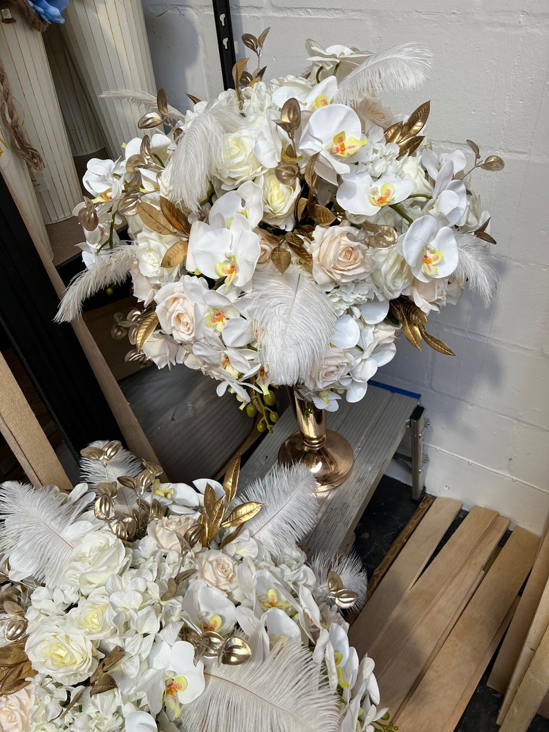 K&ouml;nnte beinhalten: Ein wei&szlig;-goldener Blumenstrau&szlig; mit wei&szlig;en Orchideen, Rosen und Hortensien. Die Anordnung befindet sich in einer goldenen Vase und ist mit wei&szlig;en Federn verziert.