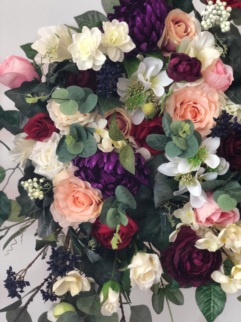 May include: A bouquet of artificial flowers in shades of pink, white, burgundy, and purple. The bouquet includes roses, hydrangeas, and eucalyptus.