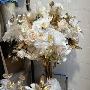 K&ouml;nnte beinhalten: Ein wei&szlig;-goldener Blumenstrau&szlig; mit wei&szlig;en Orchideen, Rosen und Federn in einer goldenen Vase.