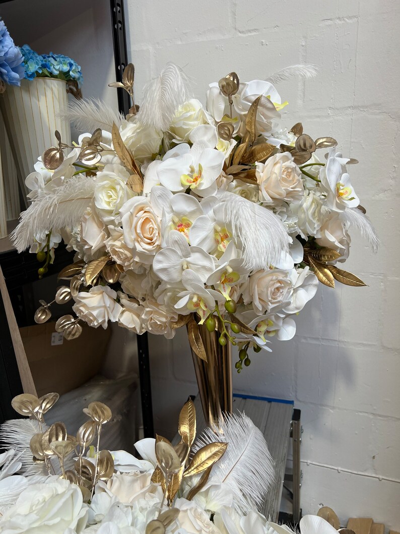 K&ouml;nnte beinhalten: Ein wei&szlig;-goldener Blumenstrau&szlig; mit wei&szlig;en Orchideen, Rosen und Federn in einer goldenen Vase.