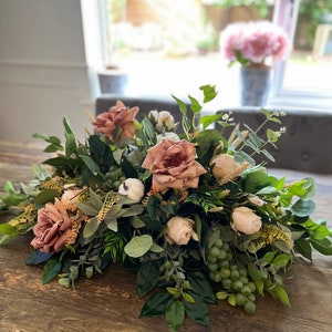 May include: A floral centerpiece featuring mauve roses, white pumpkins, and green grapes, surrounded by lush greenery. The arrangement is set on a wooden table, creating a natural and elegant display. Perfect for home decor or special events.