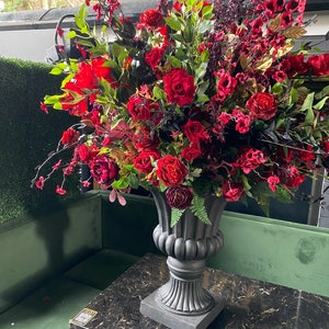 Pu&ograve; includere: Una grande composizione floreale con rose rosse e altri fiori rosso scuro in un'urna grigia ornata. La composizione &egrave; posta su un tavolo di marmo nero, con una panca di velluto verde sullo sfondo.