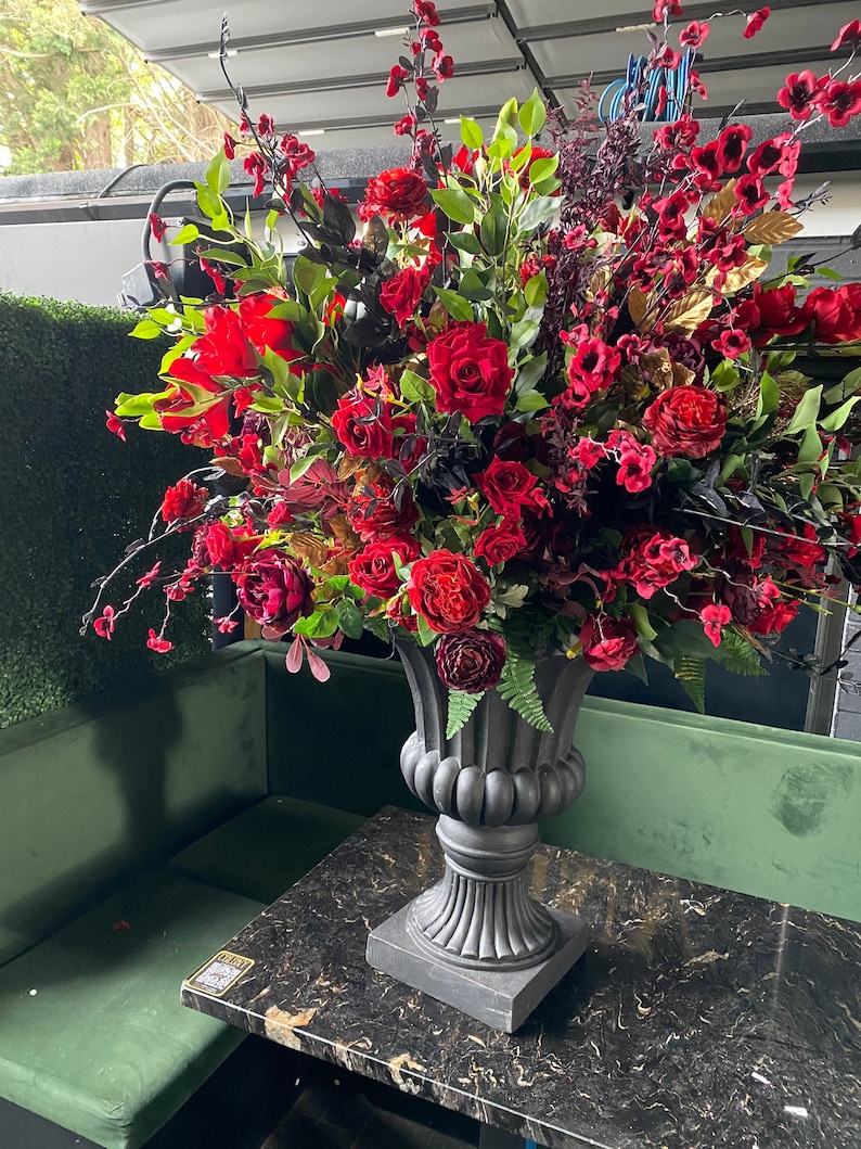 Pu&ograve; includere: Una grande composizione floreale con rose rosse e altri fiori rosso scuro in un'urna grigia ornata. La composizione &egrave; posta su un tavolo di marmo nero, con una panca di velluto verde sullo sfondo.