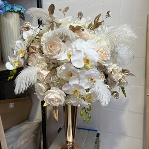 K&ouml;nnte beinhalten: Ein gro&szlig;er wei&szlig;er und goldener Blumenstrau&szlig; mit wei&szlig;en Rosen, Orchideen und Federn in einer goldenen Vase.