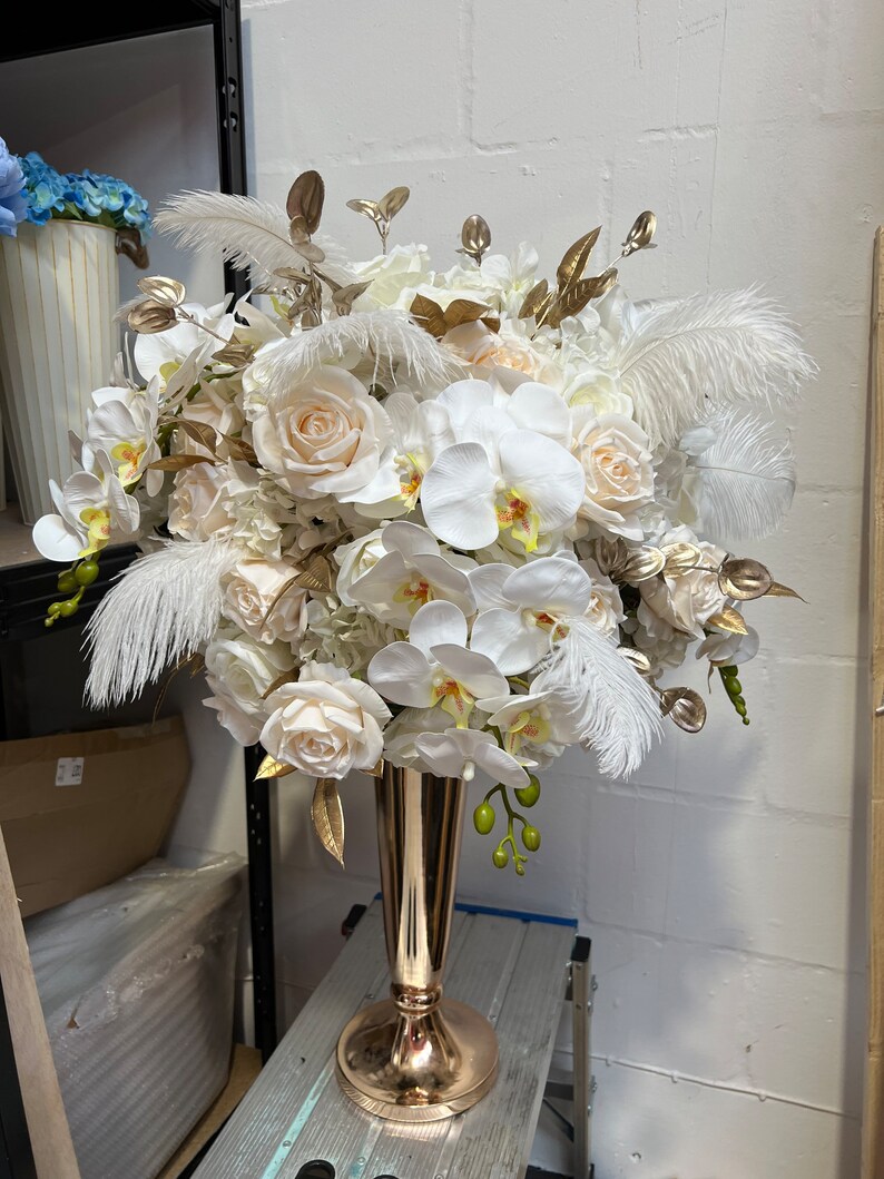 K&ouml;nnte beinhalten: Ein gro&szlig;er wei&szlig;er und goldener Blumenstrau&szlig; mit wei&szlig;en Rosen, Orchideen und Federn in einer goldenen Vase.