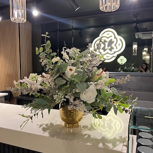 May include: A floral arrangement in a gold vase. The bouquet features white roses, green foliage, and small white flowers. The vase has a textured surface and sits on a white countertop. The background includes a dark wall and decorative lighting.