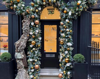 Puede incluir: Entrada de una tienda decorada para las fiestas. La puerta negra está enmarcada por una guirnalda de ramas de hoja perenne, adornos dorados y blancos y flores blancas. Un gran elemento con textura similar a un tronco de árbol se encuentra en el lado izquierdo.