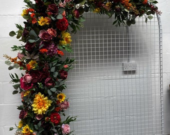 Op de afbeelding: Een bloemenboog gemaakt van kunstbloemen in tinten rood, geel en roze. De bloemen zijn in een cascade-ontwerp gerangschikt en zijn bevestigd aan een wit metalen rooster.