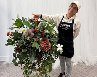 May include: A large, lush floral arrangement featuring a variety of flowers and greenery in shades of red, white, green, and brown. The arrangement includes cascading ivy and is held by a person wearing a black apron.