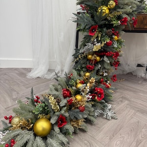 Puede incluir: Una guirnalda navideña festiva con ramas de pino artificiales, bayas rojas, adornos dorados y flores rojas. La guirnalda está dispuesta en un diseño decorativo.