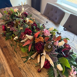 Peut inclure: Une composition florale colorée pour centre de table, avec des roses bordeaux, des tulipes roses et blanches, et divers feuillages. L'arrangement comprend des éléments décoratifs comme de petites citrouilles, sur une table en bois rustique. Les couleurs sont riches et automnales.