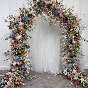May include: A decorative floral archway, featuring a profusion of colourful flowers in shades of pink, blue, yellow, and white. The arch is crafted from branches and foliage, creating a natural look. A white curtain serves as the backdrop.