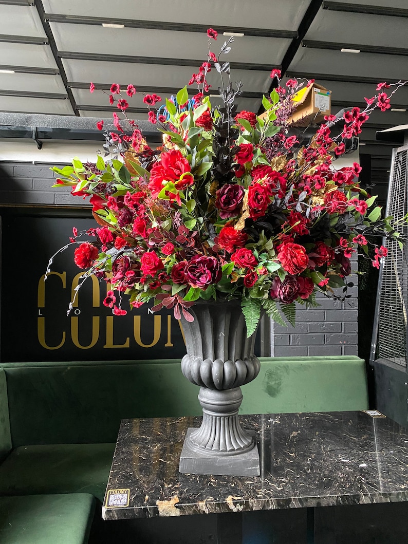 Pu&ograve; includere: Un grande vaso grigio ornato traboccante di vibranti fiori e verde rosso e bordeaux. La composizione comprende rose, peonie e altri fiori. Il vaso &egrave; posto su un tavolo con motivo marmoreo davanti a una panca di velluto verde.