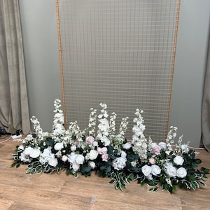 May include: A gold metal archway with a white and pink floral arrangement at the base. The flowers are arranged in a cascading style and include white roses, pink roses, and white hydrangeas.