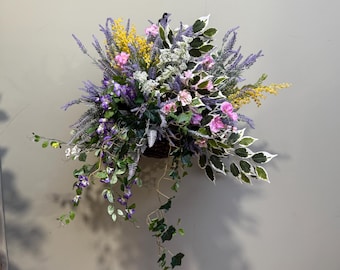 May include: A hanging flower basket filled with a variety of purple, pink, and yellow flowers. The basket is made of woven material and is hanging from a wall.