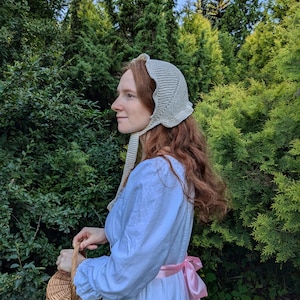 Może przedstawiać: Kobieta w białym, szydełkowym czepeczku i jasnoniebieskiej sukience z różową wstążką zawiązaną w talii. Trzyma wiklinowy koszyk w lewej ręce. Kobieta stoi w lesie.