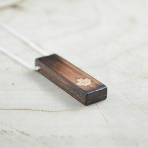 May include: A wooden pendant necklace featuring a rectangular, dark brown pendant with a maple leaf design. The pendant is suspended from a delicate silver chain. The necklace is displayed on a light-colored, textured surface.