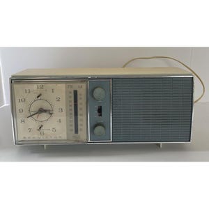 Puede incluir: Radio reloj RCA Victor vintage con una parte superior color crema y un panel de altavoz gris. La esfera del reloj es blanca con números y manecillas negras. La radio tiene dos perillas y un interruptor, con indicadores de frecuencia de radio FM y AM.