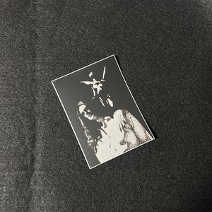 May include: A black and white sticker featuring an illustration of a person with long hair and a figure above them. The sticker is rectangular with a white border and is placed on a dark gray surface.