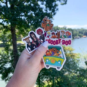 May include: A collection of colorful Scooby-Doo stickers featuring the Mystery Inc. gang and the Mystery Machine van. The stickers showcase various characters and scenes from the animated series, with vibrant colors and detailed designs.