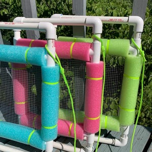 May include: Two square frames constructed from white PVC pipe, each featuring a mesh center and foam pool noodles in bright pink, turquoise, and lime green. The frames are connected by neon green cords and display printed text on the pipes.
