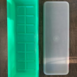 May include: A rectangular bright green plastic storage container with a grid-patterned base and a separate, translucent, rectangular lid. The lid features a circular center design and the embossed text Tupperware in the upper right corner.