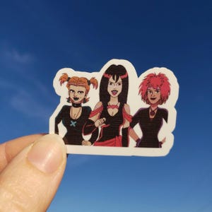 May include: A sticker featuring a cartoon illustration of three women with dark makeup and black outfits. One has orange pigtails, one has black hair, and the third has pink hair. The sticker is held against a blue sky.