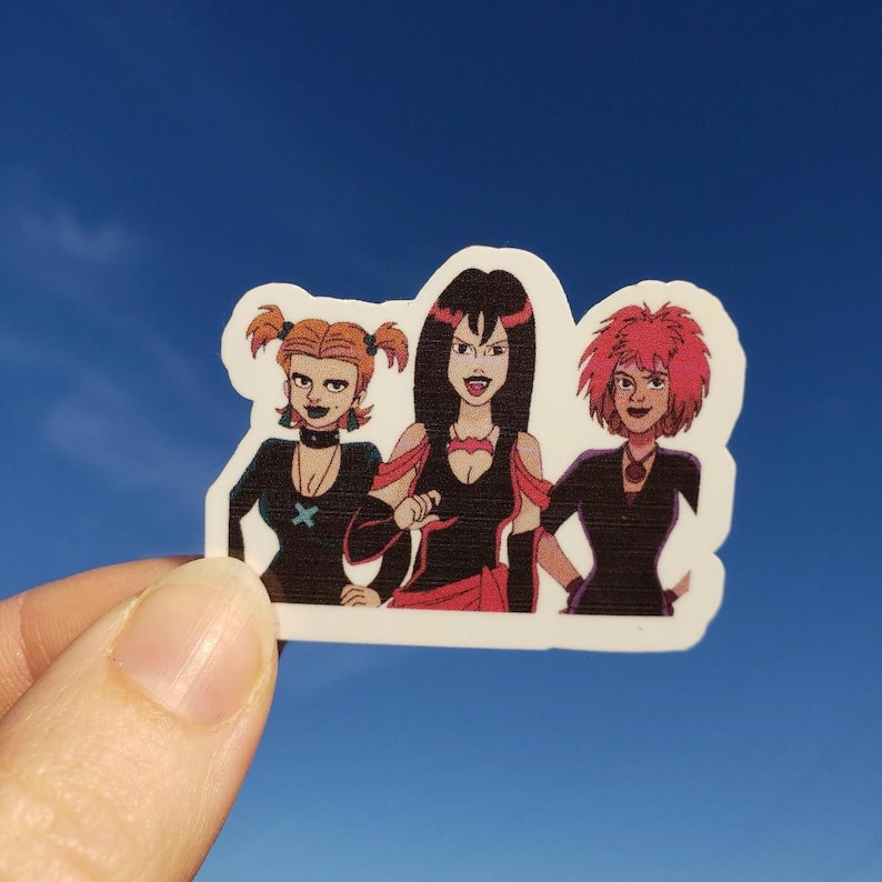 May include: A sticker featuring a cartoon illustration of three women with dark makeup and black outfits. One has orange pigtails, one has black hair, and the third has pink hair. The sticker is held against a blue sky.