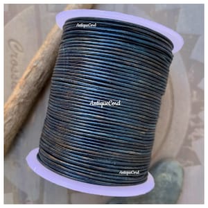 May include: A spool of black leather cord with a subtle metallic sheen. The cord is wound tightly around a white plastic spool. The text "AntiqueCord" is printed on the spool.