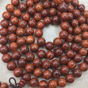 May include: A strand of dark red wooden beads, likely made of rosewood, with a knotted tassel at one end. The beads are round and smooth, with a natural wood grain pattern.