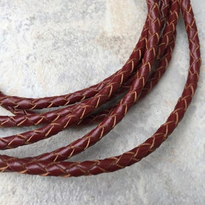 May include: Close-up of a braided leather cord in a deep burgundy color. The cord is woven with a lighter brown thread, creating a contrasting effect.