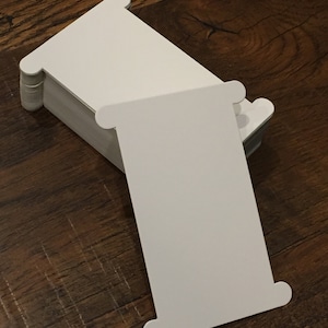 May include: A stack of white rectangular cards with rounded ends, resting on a wooden surface. The cards are blank and appear to be unused. The design features a unique shape, suitable for various creative projects.