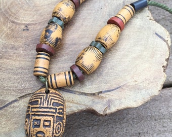 May include: A necklace made of brown, black, and green beads with a carved wooden pendant featuring a face. The pendant is oval shaped and has a detailed design.